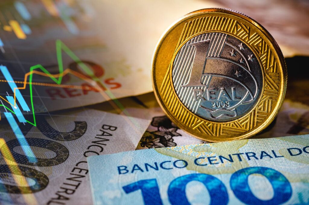 Moeda de um real destacada sobre cédulas de 100 e 200 reais, ilustrando o impacto das novas alíquotas de IOF para empresas e o aumento do custo do crédito no Brasil.